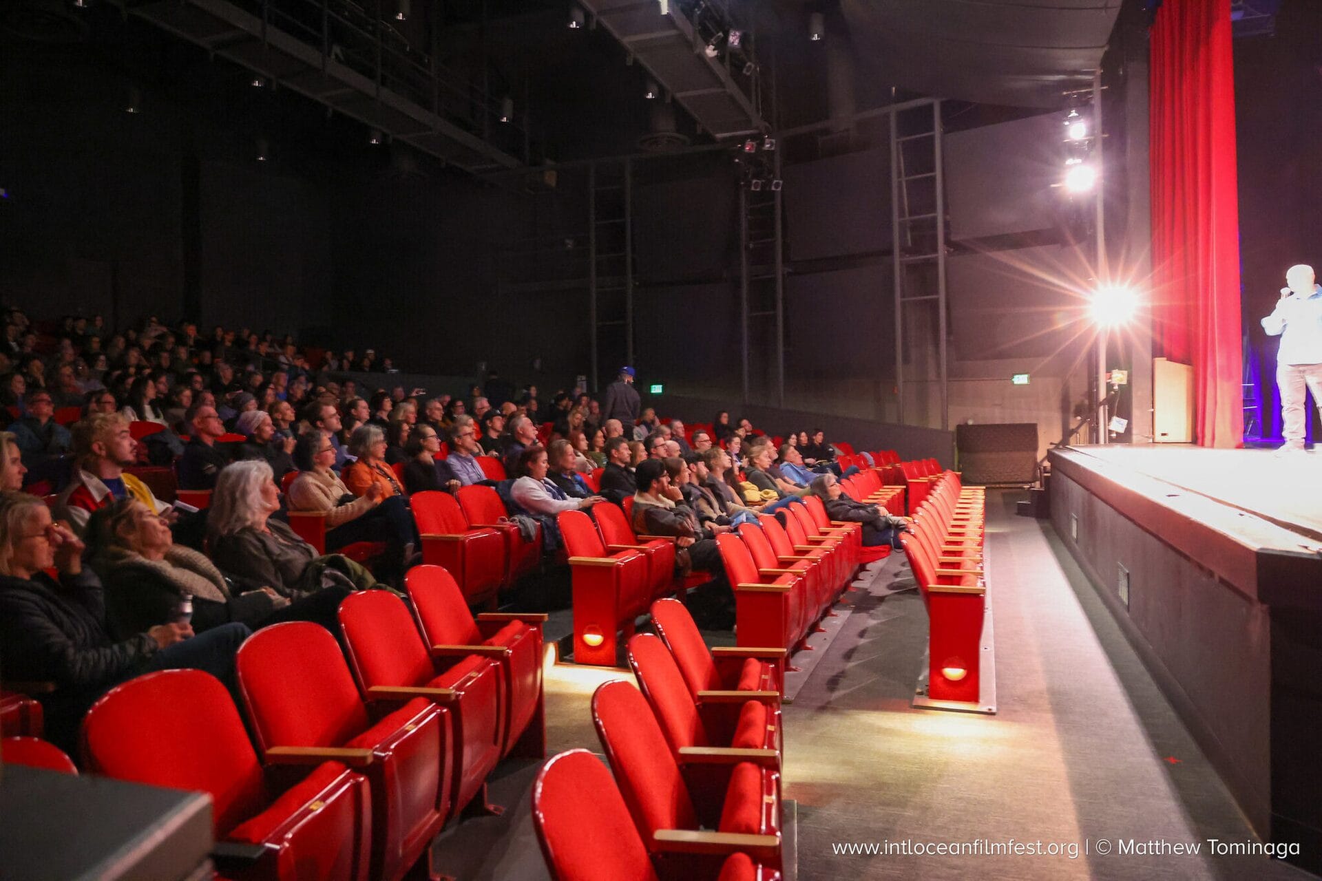 Audience filling a theater at the International Ocean Film Festival as Andi Cross presents on the lit stage
